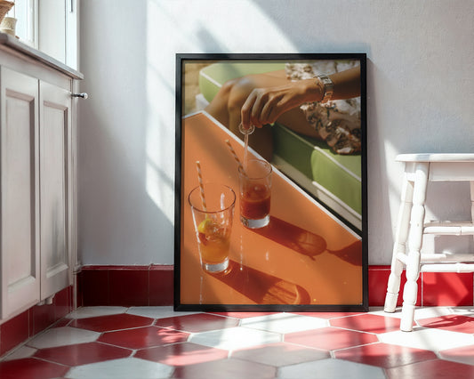A framed art print leaning against a white wall on a red and white hexagonal tile floor. The print shows a close-up photograph of a hand pouring a drink into a glass on an orange table, with a green sofa in the background.