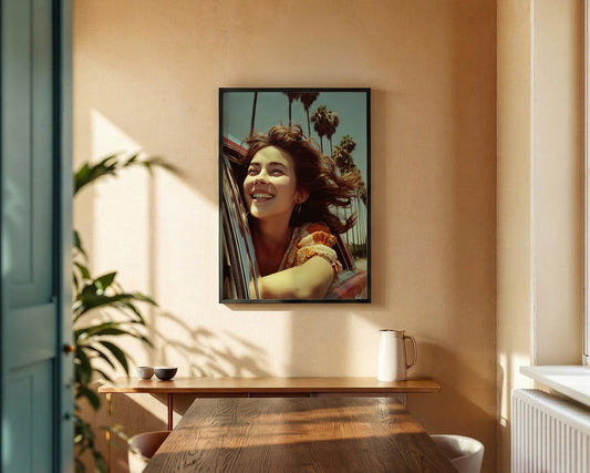A framed vertical art print featuring a smiling woman leaning out of a car with palm trees in the background, displayed on a warm beige wall above a wooden console table in a sunlit room.