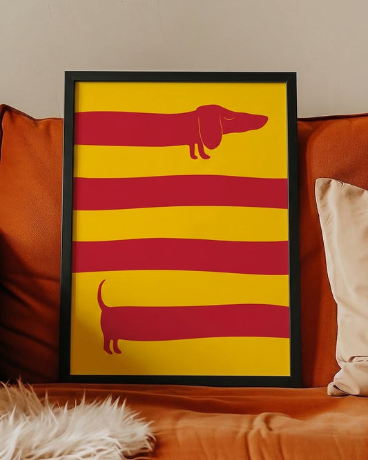 A framed art print featuring two red dachshund silhouettes on a yellow background with horizontal red stripes, leaning against an orange couch with pillows.