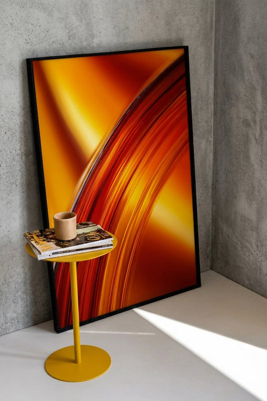 A large framed abstract art print with sweeping orange, yellow, and red brushstrokes leans against a gray concrete wall on a white floor, next to a small yellow side table holding a mug and books.