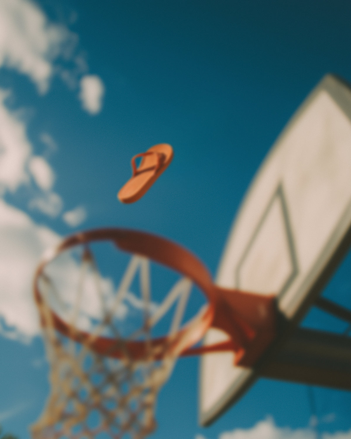 An orange flip-flop sandal floating in mid-air above a basketball hoop, set against a bright blue sky with white clouds and a basketball backboard.
