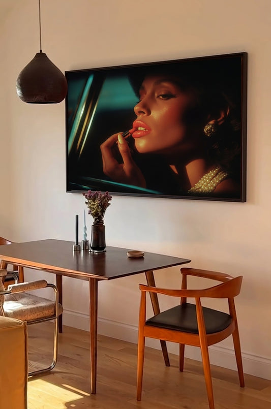 A large framed wall art print depicting a close-up portrait of a woman applying lipstick, hung above a dining table in a modern room with chairs and a pendant light.