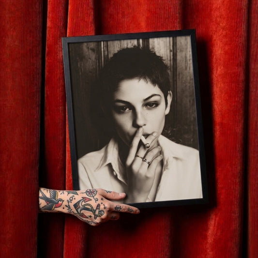 A framed black and white portrait print of a person with short dark hair, held by a tattooed hand in front of a red velvet curtain.