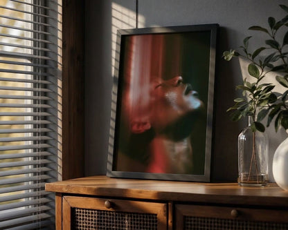 A framed vertical art print leaning on a wooden cabinet in a styled room. The print features an abstract portrait of a person's face with dramatic red and dark green tones.