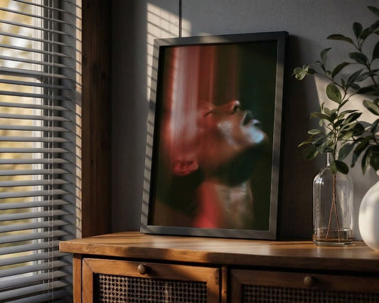 A framed vertical art print leaning on a wooden cabinet in a styled room. The print features an abstract portrait of a person's face with dramatic red and dark green tones.