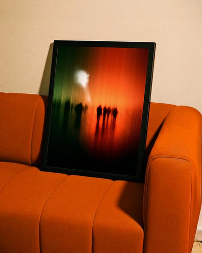 A framed abstract art print leaning against an orange sofa against a beige wall. The print features a black frame and an abstract design with a green-to-red gradient and silhouetted figures.