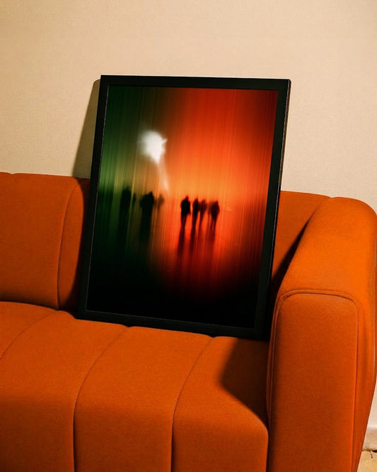A framed abstract art print leaning against an orange sofa against a beige wall. The print features a black frame and an abstract design with a green-to-red gradient and silhouetted figures.