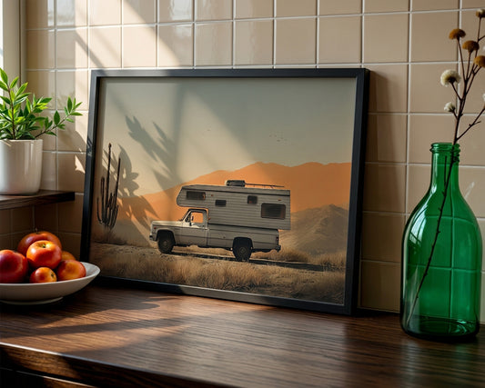 A framed art print leaning against a beige tiled wall on a wooden counter. The print shows a vintage pickup truck with a camper trailer driving through a desert landscape at sunset.
