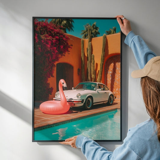 A framed art print depicting a classic white sports car parked by a turquoise pool next to a pink inflatable flamingo, with cacti and a warm orange stucco wall in the background. The print is being held up against a white wall by a person wearing a light blue sweatshirt and a beige cap.