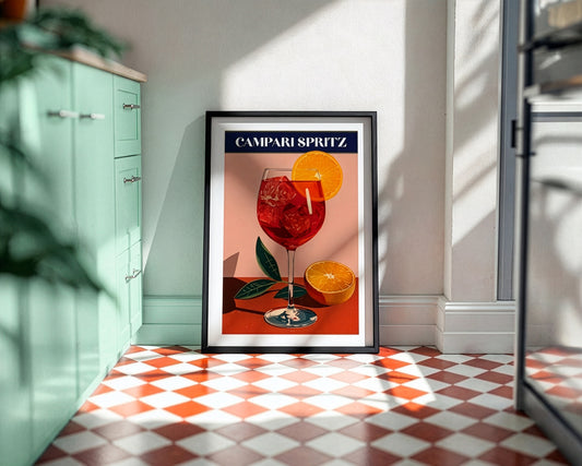 A framed art print of a vintage Campari Spritz cocktail poster leaning against a wall in a modern kitchen setting with a red and white checkered floor.