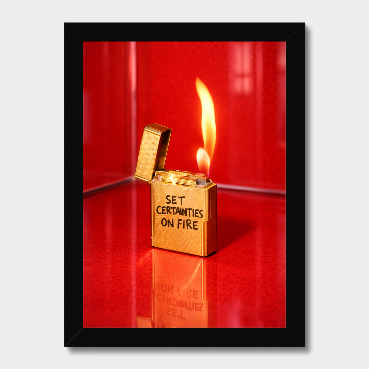 A framed photographic print featuring a gold lighter with the text 'SET CERTANTIES ON FIRE' and an open flame, set against a vivid red background, displayed on a light gray wall.
