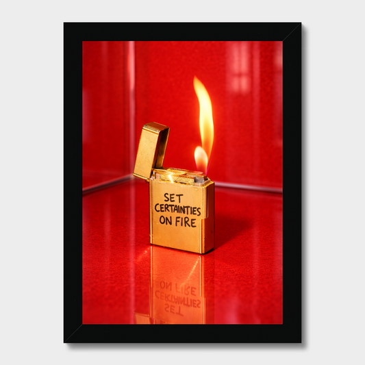 A framed photographic print featuring a gold lighter with the text 'SET CERTANTIES ON FIRE' and an open flame, set against a vivid red background, displayed on a light gray wall.
