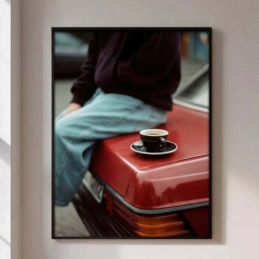 A framed vertical art print hanging on a light beige wall. The print shows a person in a dark sweater and light blue jeans sitting on the trunk of a red car, with a small black coffee cup on a saucer resting on the car.