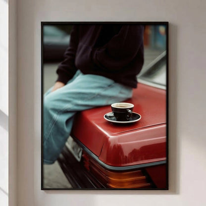 A framed vertical art print hanging on a light beige wall. The print shows a person in a dark sweater and light blue jeans sitting on the trunk of a red car, with a small black coffee cup on a saucer resting on the car.