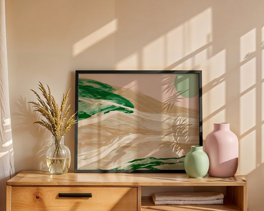 A framed abstract landscape art print with green and beige brushstrokes, styled on a wooden console table next to a glass vase with dried grass and two ceramic vases against a beige wall with window shadow patterns.