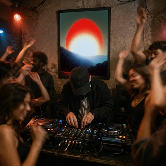 A framed wall art print depicting a large red and white sun over dark mountains hangs on a textured wall in a dimly lit club or party space, with a DJ in a black cap and jacket operating turntables while people dance around.