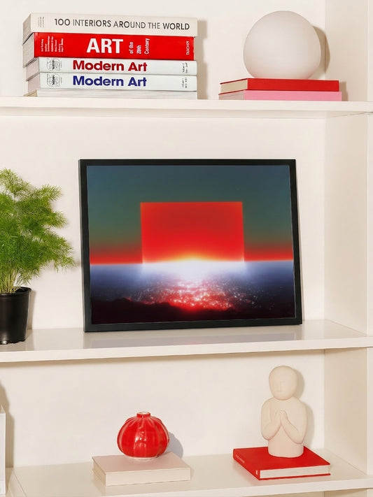 A framed abstract art print featuring a red square over a dark landscape with a bright sunburst, displayed on a white shelf alongside decorative books, a plant, and a small sculpture.