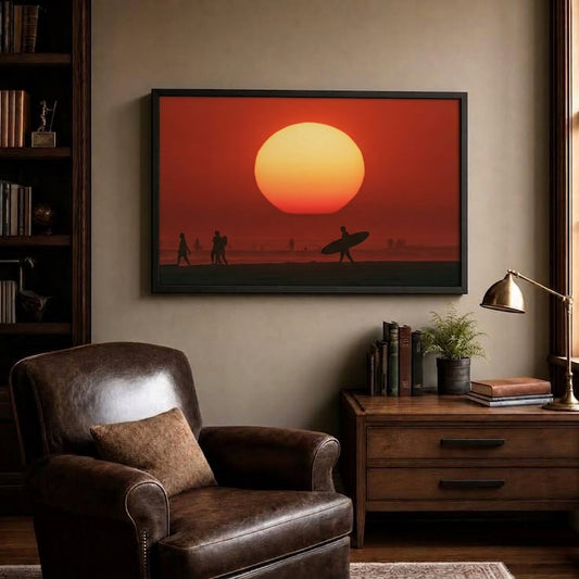 A framed wall art print showing a large orange and yellow sun over a beach silhouette scene, displayed above a brown leather armchair in a styled living room with a wooden side table and lamp.