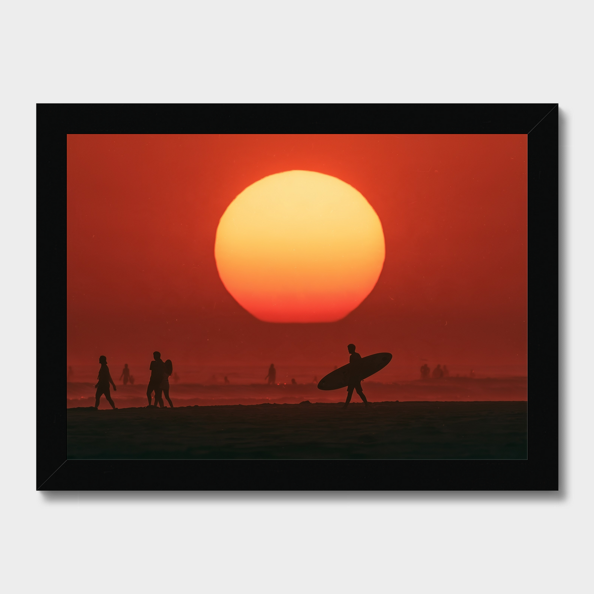 Framed wall art showing a large orange and red sunset over a beach, with silhouetted figures and a surfer carrying a board. The black frame is visible against a light gray background.