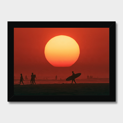 Framed wall art showing a large orange and red sunset over a beach, with silhouetted figures and a surfer carrying a board. The black frame is visible against a light gray background.