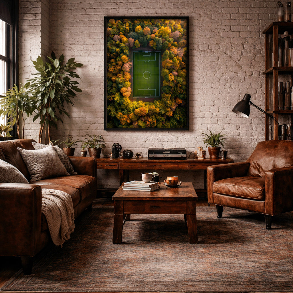 A framed wall art print depicting a green soccer field surrounded by a dense forest of yellow, orange, and green autumn trees, displayed in a cozy, modern living room with a brown leather sofa, wooden coffee table, and brick wall.