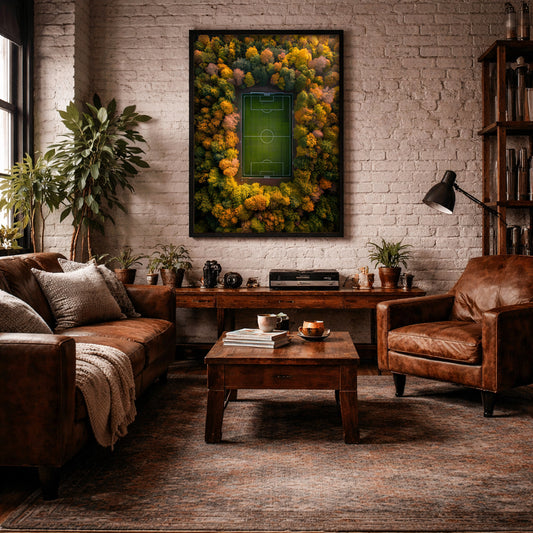 A framed wall art print depicting a green soccer field surrounded by a dense forest of yellow, orange, and green autumn trees, displayed in a cozy, modern living room with a brown leather sofa, wooden coffee table, and brick wall.