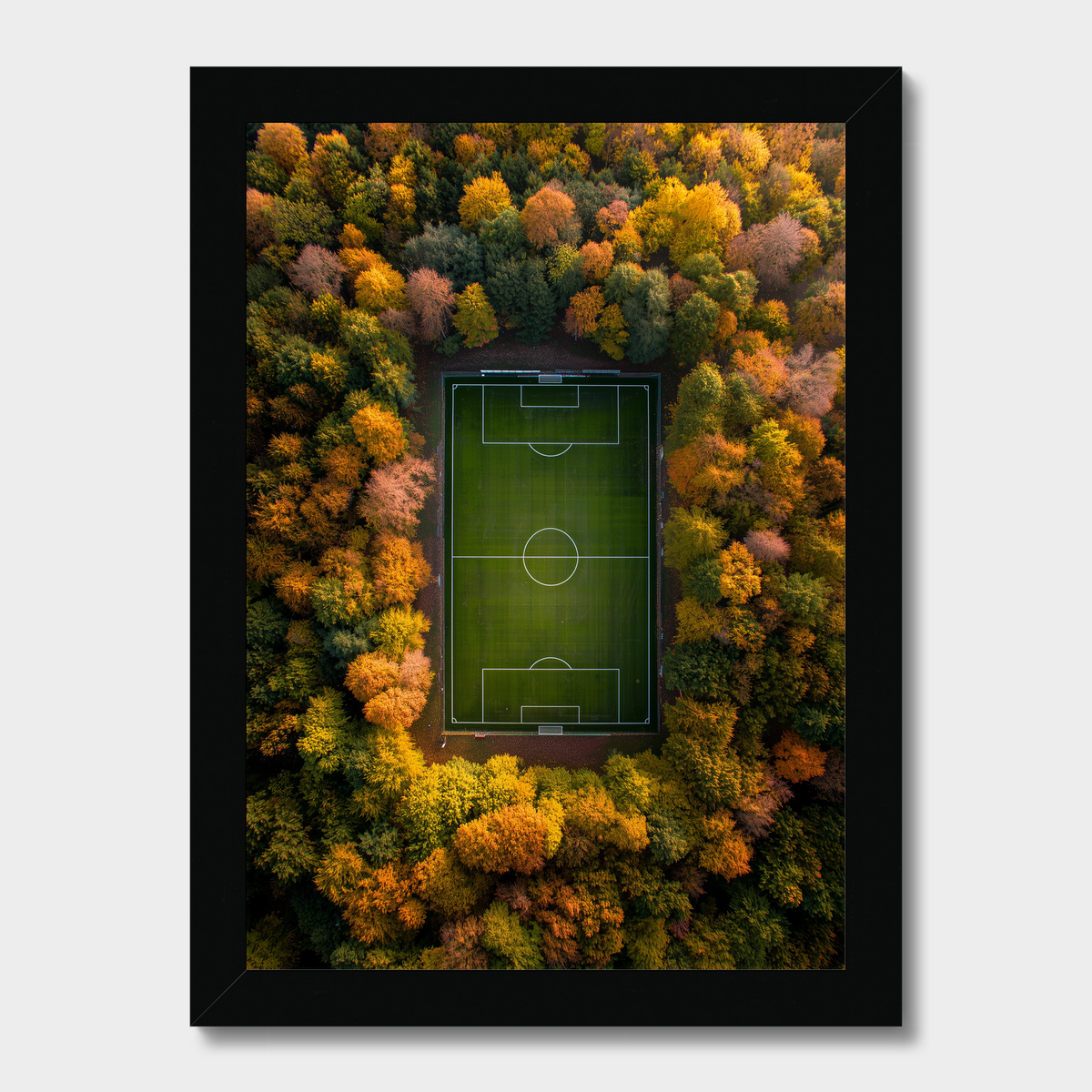 Framed wall art featuring an aerial view of a green soccer field surrounded by dense autumn forest in shades of orange, yellow, and green, shown in a black frame against a light gray background.
