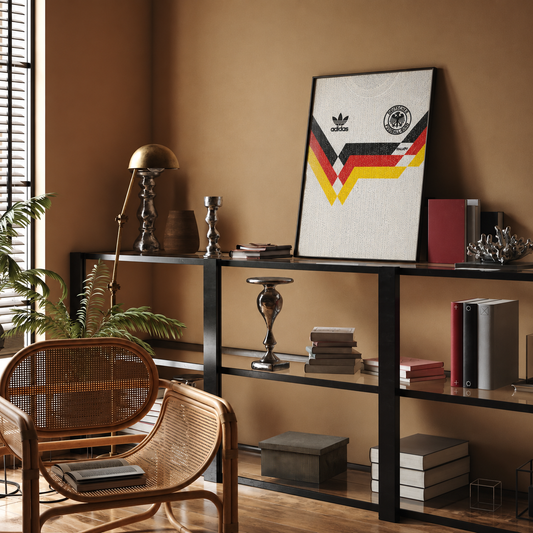 A framed Adidas Germany soccer jersey print hangs on a warm brown wall in a styled room with a rattan chair, shelf, books, and decor.