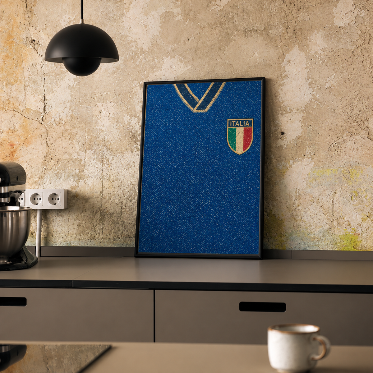 A framed blue Italy soccer jersey print leaning against a textured beige wall on a dark countertop, with a black pendant lamp, stand mixer, and white mug nearby.