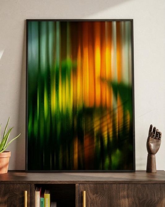 A framed abstract art print featuring vertical streaks of green, yellow, and orange, displayed on a dark wooden console table next to a small potted plant and a decorative hand sculpture.