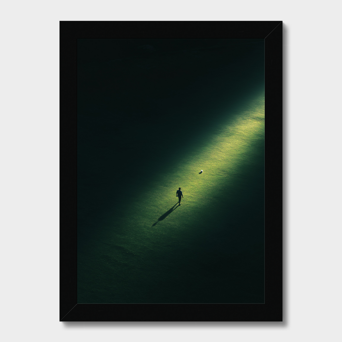 A framed art print featuring a minimalist aerial photograph of a person walking on a dark green field under a spotlight, set in a sleek black frame against a light gray background.