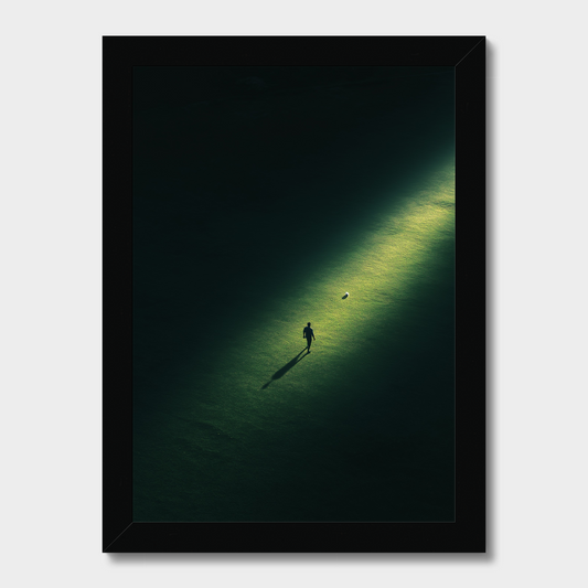 A framed art print featuring a minimalist aerial photograph of a person walking on a dark green field under a spotlight, set in a sleek black frame against a light gray background.