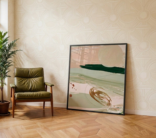 A large framed abstract art print with soft beige, sage green, and dark teal brushstrokes leans against a patterned wallpaper wall in a modern room next to an olive green leather armchair and a potted plant.