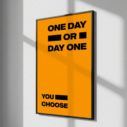 A framed motivational poster with a bright orange background and bold black text reading 'ONE DAY OR DAY ONE' and 'YOU CHOOSE', hanging on a light gray wall with soft window shadows.