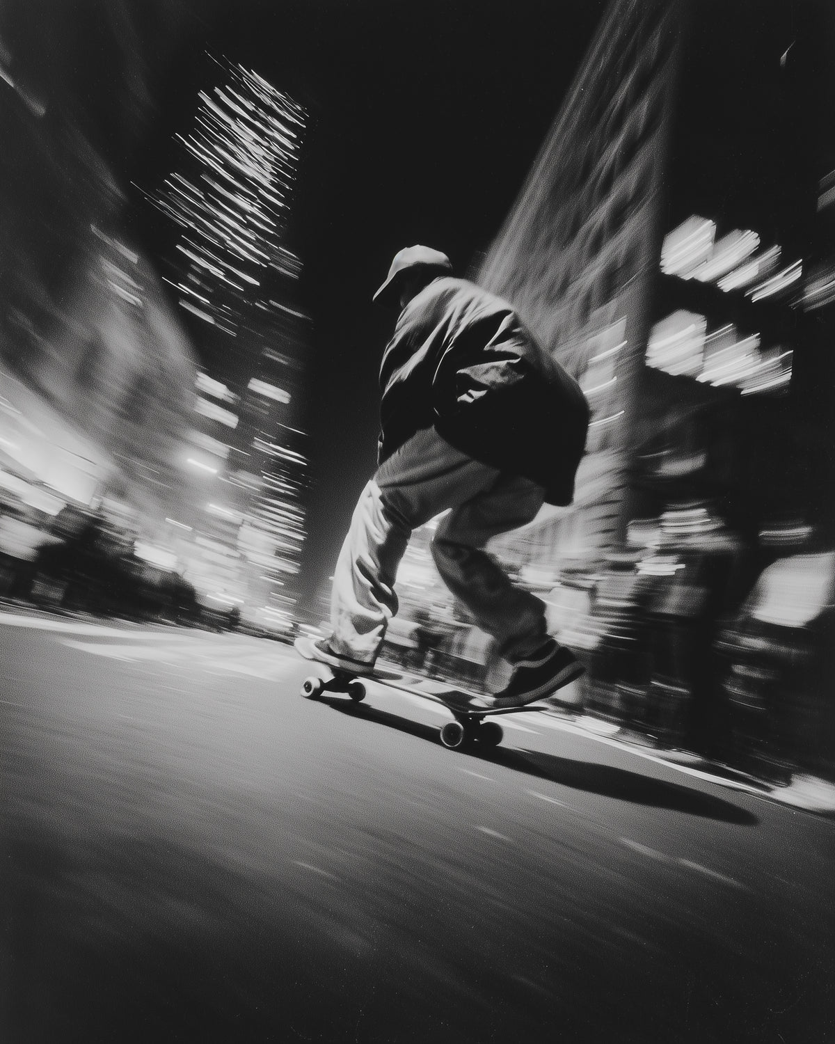 Black and white photograph of a person skateboarding on a city street at night, captured with motion blur and streaked light from surrounding buildings.