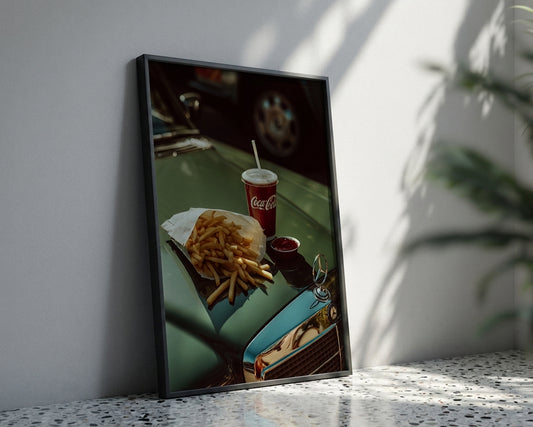 A framed vertical art print leaning against a white wall on a speckled countertop. The print shows a close-up diner-style scene with french fries, a Coca-Cola cup, and a small red dish on a teal car hood.