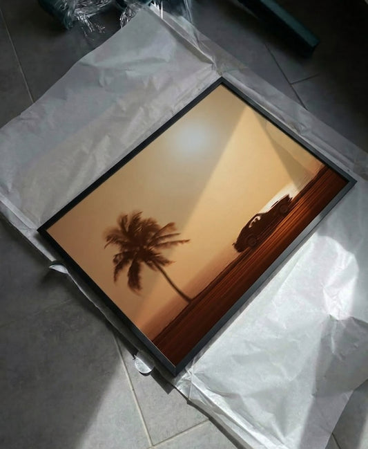 A framed art print featuring a sunset landscape with a palm tree and a car silhouette, resting on white protective paper over a gray tiled floor.