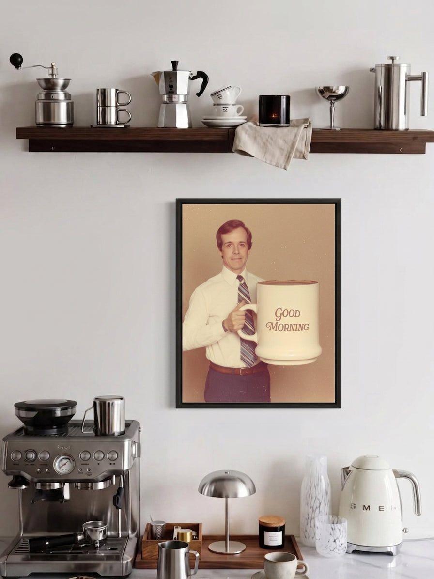 A framed vintage-style coffee-themed wall print hangs on a white wall above a countertop with coffee equipment. The print shows a man holding a large coffee mug with the words 'Good Morning' on a beige background with a black frame.