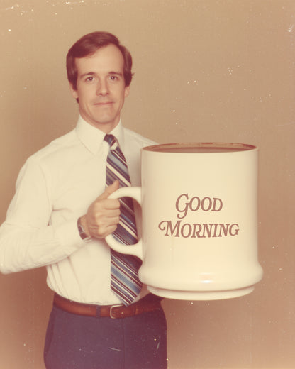 A vintage-style photo of a man in a white dress shirt and striped tie holding a very large white ceramic mug that reads 'Good Morning' in brown lettering.