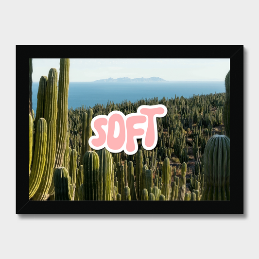 A framed art print featuring a desert landscape with green saguaro cacti and the ocean in the background, with the word 'SOFT' in large pink bubble letters overlaid in the center. The print is housed in a black frame and displayed against a light gray background.
