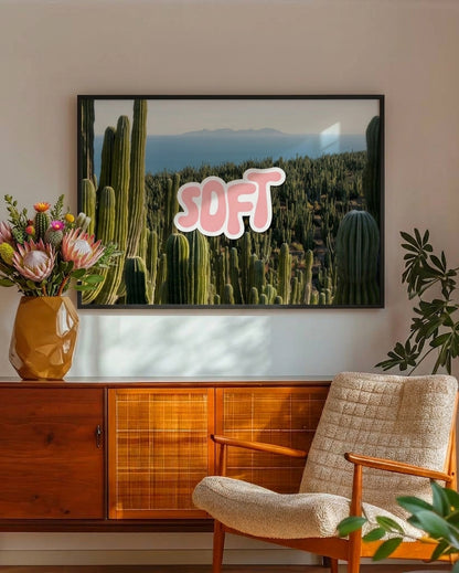 A framed wall art print featuring a desert landscape with tall green cacti and the word 'SOFT' in pink bubble letters, displayed above a wooden cabinet in a styled interior with a chair and vase of flowers.