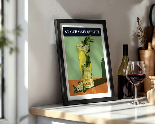 A framed vintage-style cocktail poster reading 'ST GERMAIN SPRITZ' leans on a tabletop beside a bottle of red wine and a filled wine glass in a sunlit interior.