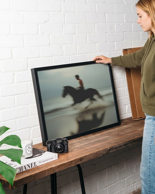 A woman in an olive green hoodie and blue jeans holds up a large black-framed art print leaning on a rustic wooden table against a white brick wall. The print shows a blurred photo of a person riding a horse on a beach.