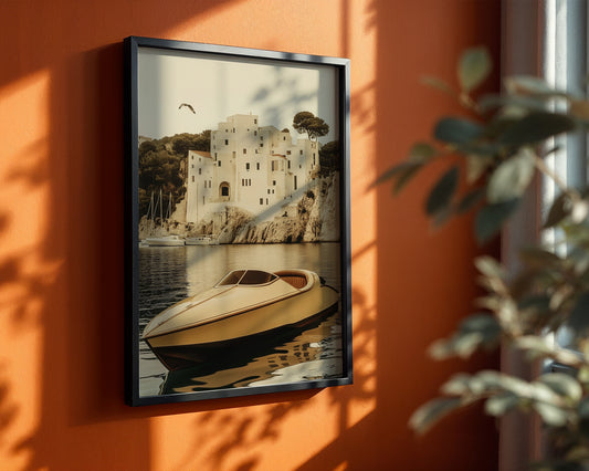 A framed art print depicting a white villa on a rocky coast with a speedboat on the water, hanging on a warm orange wall with natural shadows and a blurred plant in the foreground.
