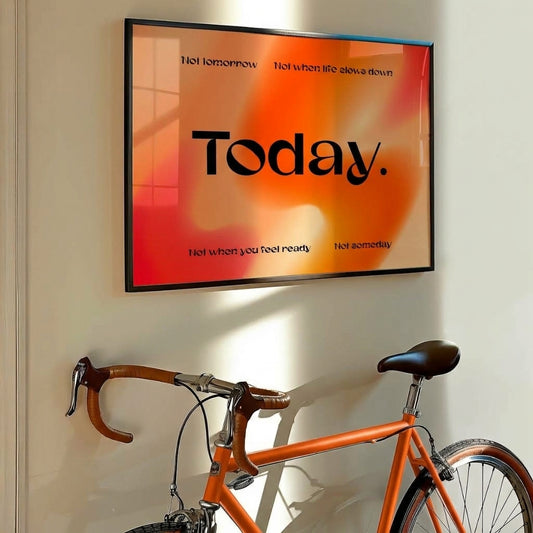 A framed wall art print with the word 'Today.' in large black text over a warm orange and red gradient background, hanging on a beige wall above an orange bicycle.