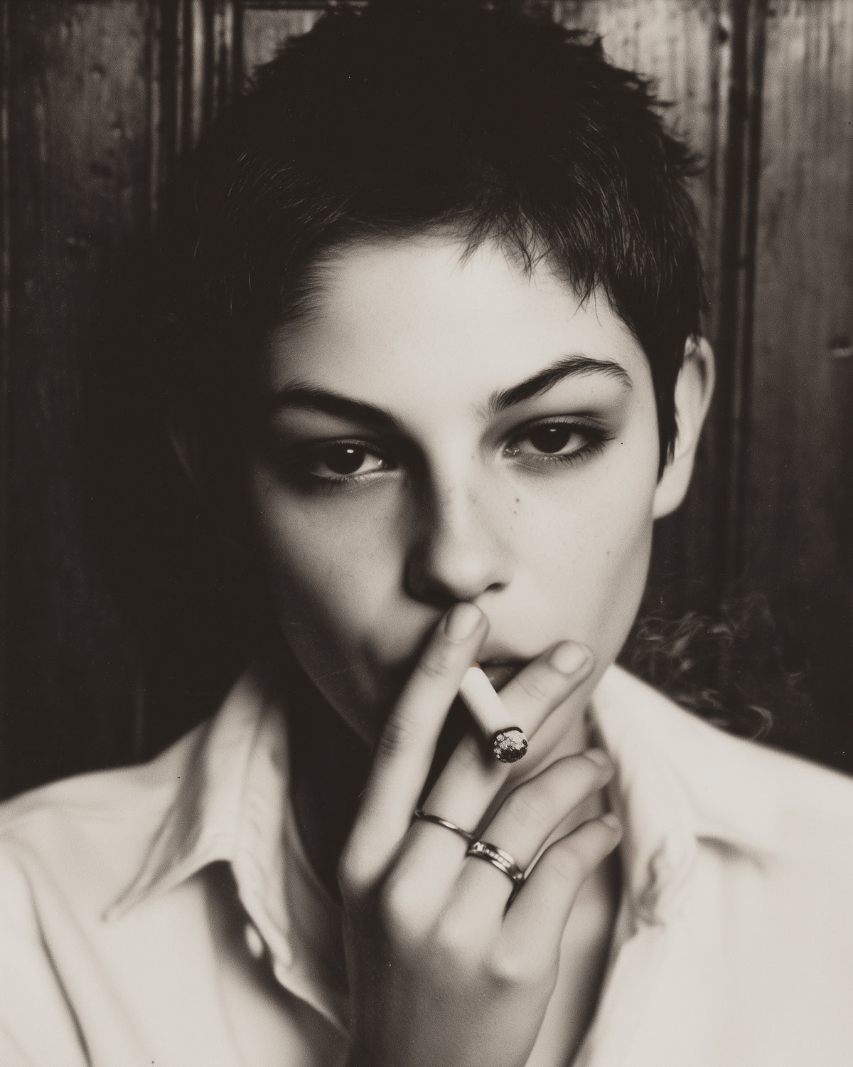 A black and white portrait of a person with short hair, wearing a light-colored collared shirt and multiple rings, holding a cigarette near their lips against a dark wood background.
